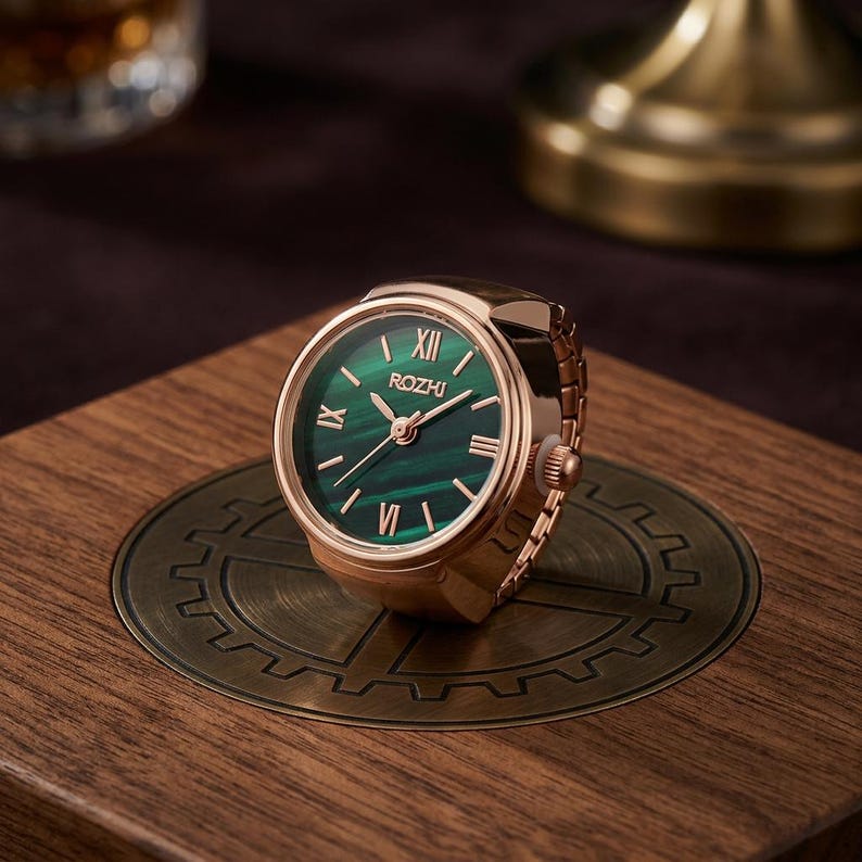 May include: A gold-toned ring watch with a green face and Roman numeral hour markers. The watch face displays the brand name "ROZHI". The watch is set on a wooden surface with a decorative metal plate.