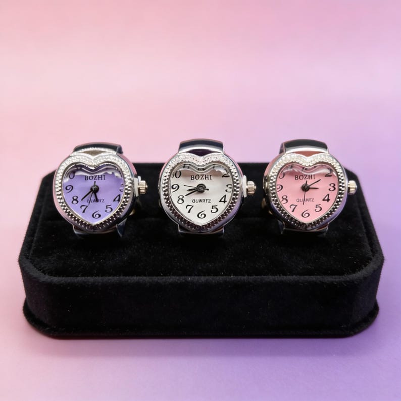 May include: Three heart-shaped quartz ring watches with silver metal bands. The watch faces display the brand "BOZHI" and feature purple, white, and pink backgrounds. The rings are displayed on a black velvet surface against a pink and purple backdrop.