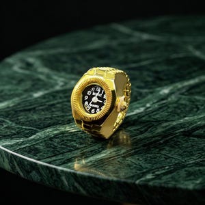 May include: A gold-colored ring watch with a black watch face. The watch face displays white numerals and hands, with the brand name "BOBHI" at the top. The ring is set on a green marble surface.