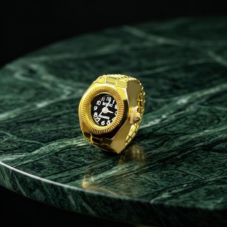 May include: A gold-colored ring watch with a black watch face. The watch face displays white numerals and hands, with the brand name "BOBHI" at the top. The ring is set on a green marble surface.