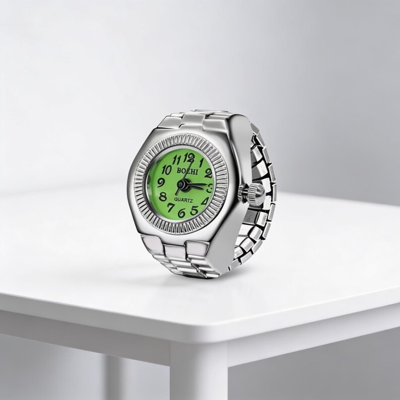 May include: A silver ring watch with a green face and quartz movement. The watch face is round with black numerals and hands. The band is made of linked metal. The watch is displayed on a white surface.