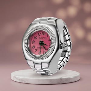 May include: A silver ring watch with a pink face and black hands, displayed on a white marble stand. The watch face shows the numbers 1 to 12 and the word "BOZHI". The watch band is made of silver links.