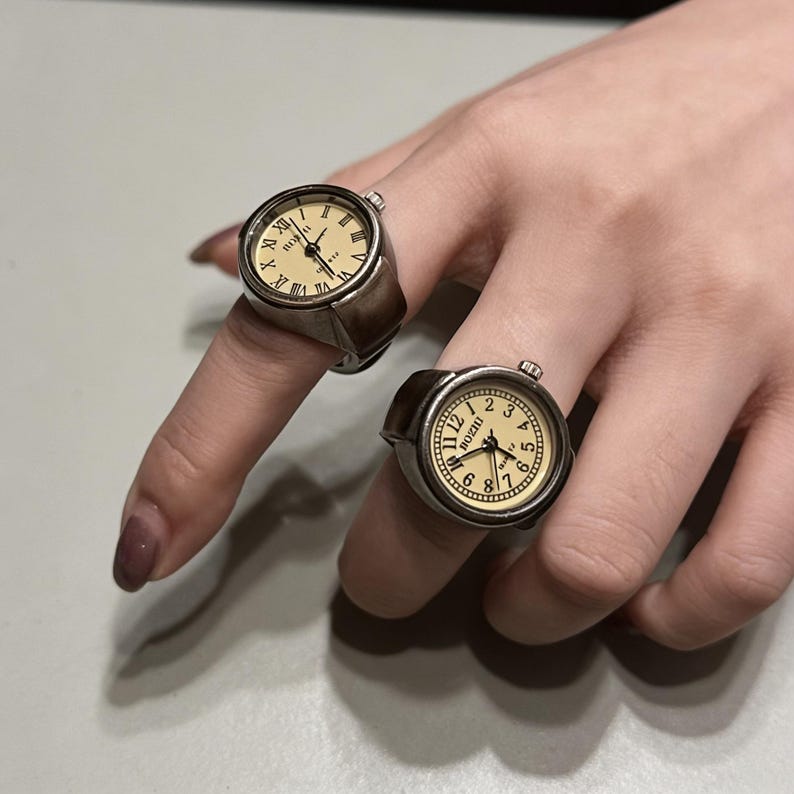 May include: Two silver-toned watch rings with cream-colored faces and black Roman numeral hour markers. The rings have a vintage aesthetic, with the watch faces displaying the time. The rings are worn on fingers.