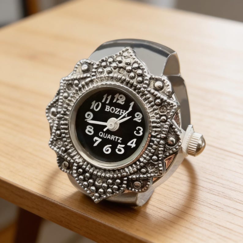May include: A silver ring watch with an ornate, floral-inspired bezel. The watch face is black with white numerals and hands, and the word "QUARTZ" is printed at the bottom. The watch is resting on a wooden surface.