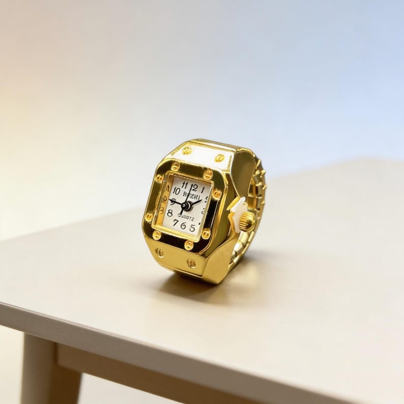 May include: A gold-colored ring watch with a square face and a white dial. The watch has black numerals and hands, and the band is made of small, connected gold links. The watch is sitting on a light-colored surface.