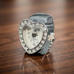 May include: A silver heart-shaped ring watch with a white face and crystal accents. The watch face displays the time with black numerals and hands. The band is silver and has a small knob on the side.