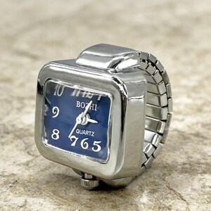 May include: A silver-toned ring watch with a square face and a blue dial. The watch face displays the time with white numerals and hands. The band is a silver, expandable style. The watch face reads "BOZHI QUARTZ".