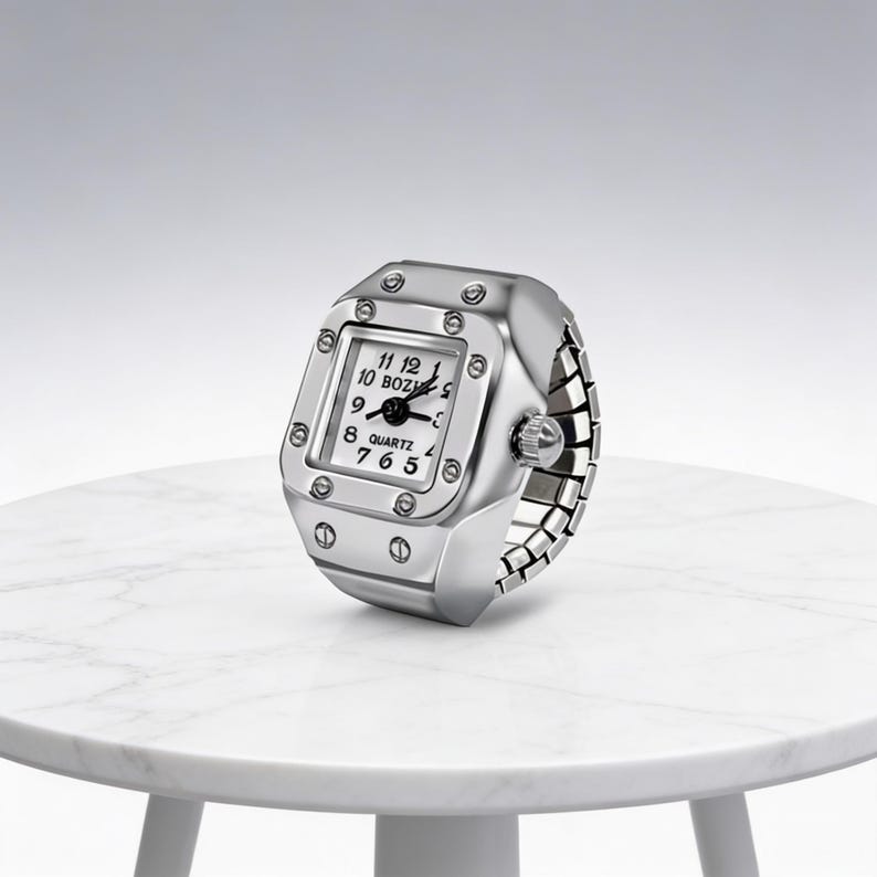 May include: A silver-toned quartz ring watch with a square face and a flexible band. The watch face displays the time with black hands and numerals. The ring watch is on a white marble table.