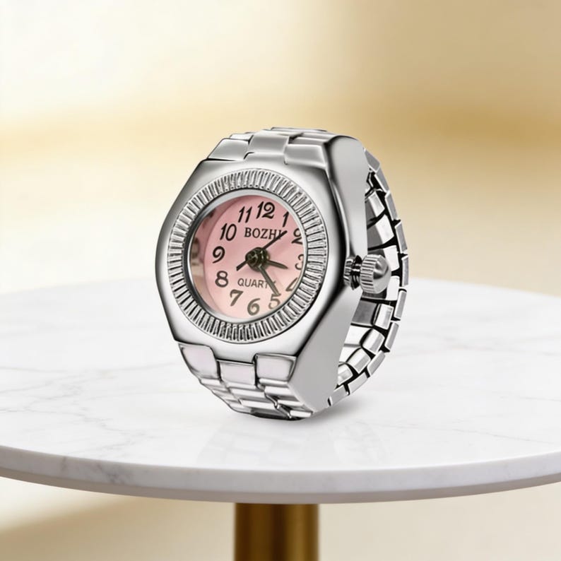 May include: A silver ring watch with a pink face and black numerals. The watch face has the word "BOZHY" printed on it. The band is made of linked metal pieces. The watch is sitting on a white marble surface.