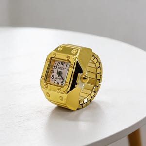 May include: A gold-colored ring watch with a rectangular face and a stretch band. The watch face displays the time with black numerals and hands. The watch is on a white surface.