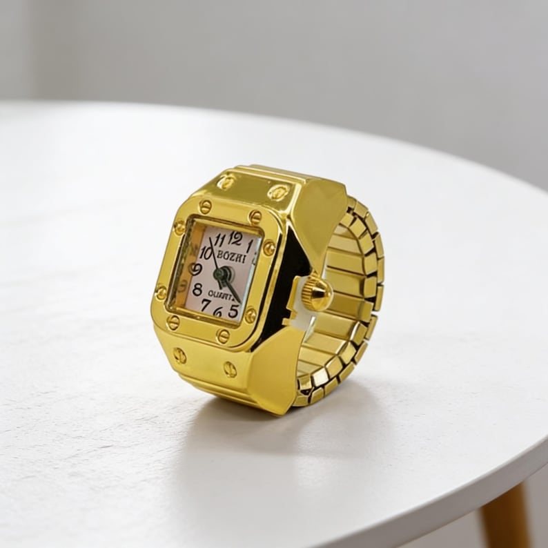 May include: A gold-colored ring watch with a rectangular face and a stretch band. The watch face displays the time with black numerals and hands. The watch is on a white surface.