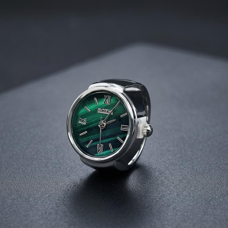 May include: A silver ring watch with a green face. The watch face has Roman numerals and the brand name "BOZH". The ring watch is set against a dark background.