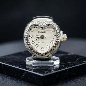 May include: A silver ring watch with a heart-shaped face. The watch face is white with black numerals and hands, and the word "BOZHI" is printed at the top. The watch is set on a clear, hexagonal base.