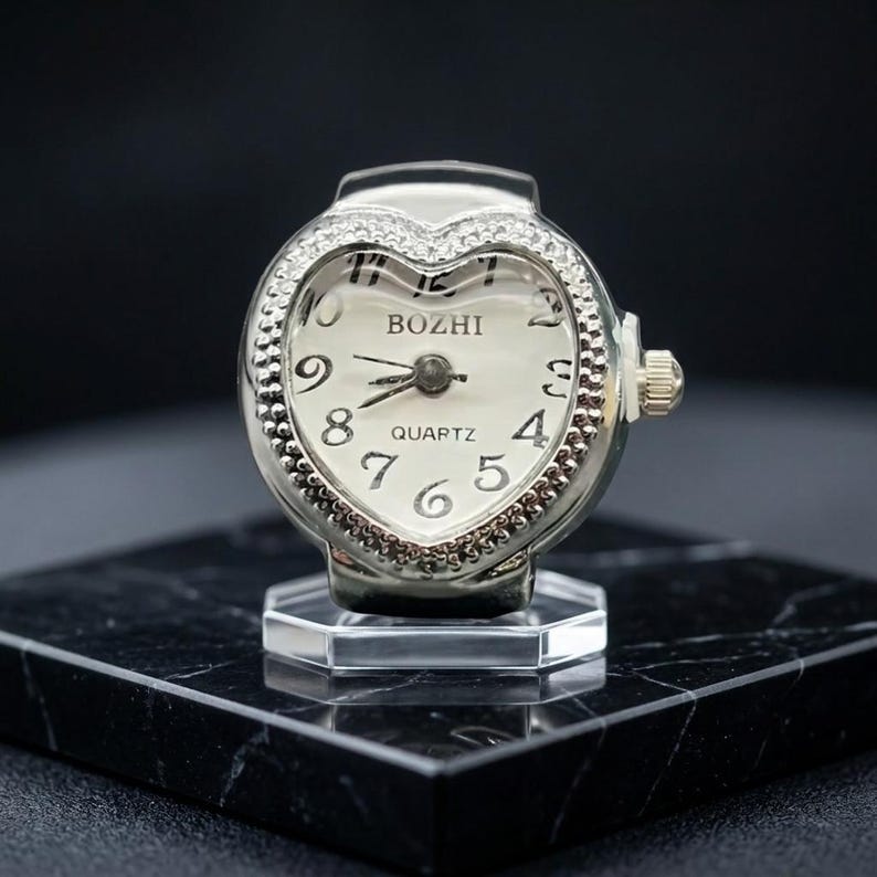 May include: A silver ring watch with a heart-shaped face. The watch face is white with black numerals and hands, and the word "BOZHI" is printed at the top. The watch is set on a clear, hexagonal base.