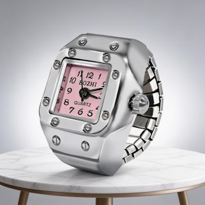 May include: A silver-toned ring watch with a square pink face. The watch face displays black numerals and hands, with the word "BOZHI" printed above the center. The watch is on a white marble table.