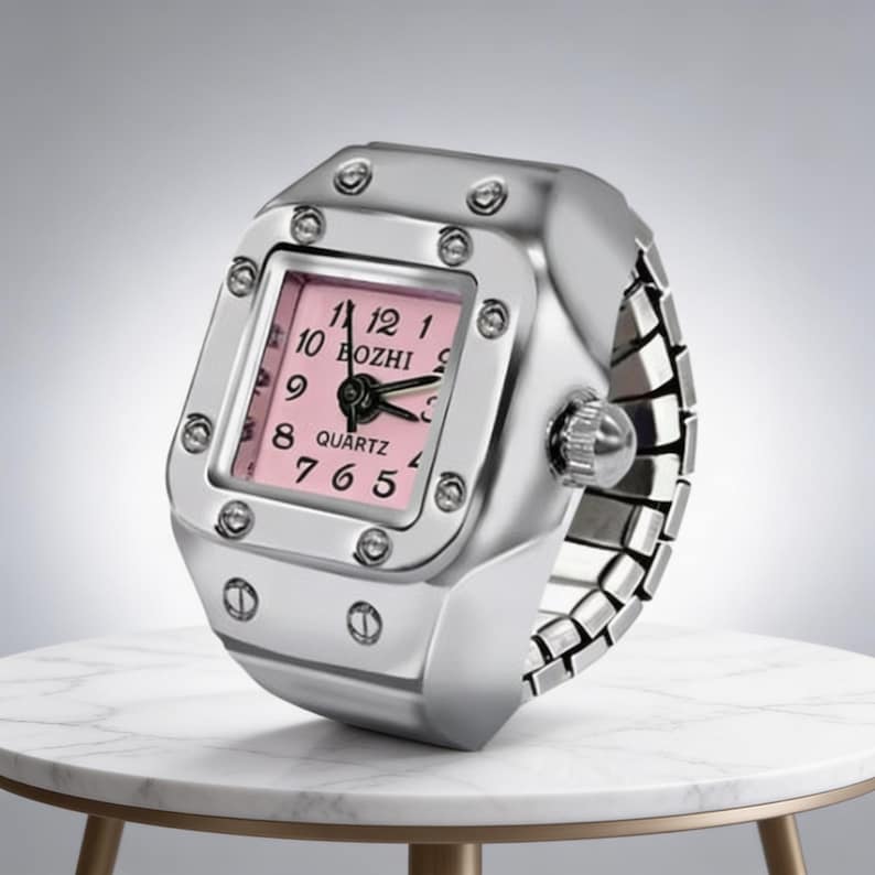 May include: A silver-toned ring watch with a square pink face. The watch face displays black numerals and hands, with the word "BOZHI" printed above the center. The watch is on a white marble table.