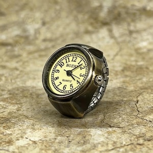 May include: Antique bronze-colored ring watch with a cream-colored face. The watch face displays black numerals and hands, with the word "QUARTZ" at the bottom. The ring has an adjustable band and a classic, vintage aesthetic.