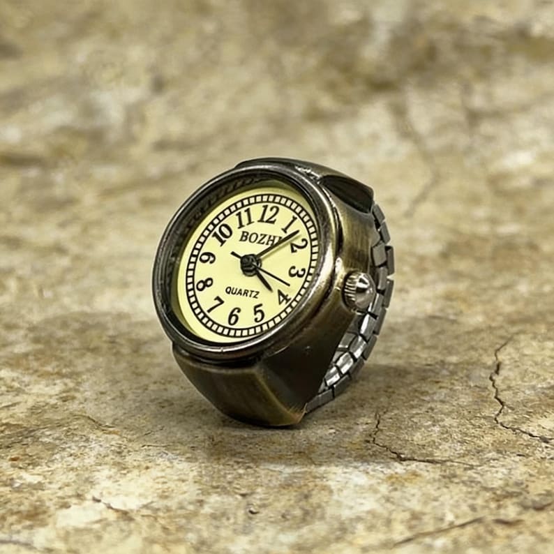 May include: Antique bronze-colored ring watch with a cream-colored face. The watch face displays black numerals and hands, with the word "QUARTZ" at the bottom. The ring has an adjustable band and a classic, vintage aesthetic.