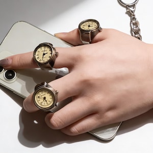 May include: Three antique-style watch rings with cream-colored faces and black numerals are displayed on a hand. The rings have a bronze-colored metal finish. The watch rings are on a white surface.