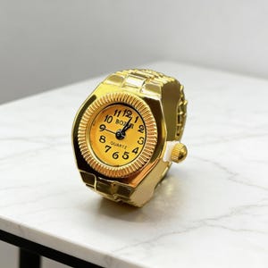 May include: A gold-colored ring watch with a round, yellow face. The watch has black numerals and hands, and the word "QUARTZ" is printed on the face. The ring band is made of gold-colored metal.