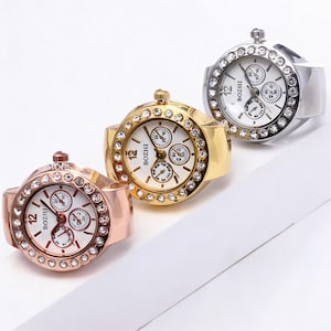 May include: Three ring watches in rose gold, gold, and silver. Each ring watch features a white face with black numerals and hands, and three smaller dials. The rings are adorned with clear crystals around the watch face.