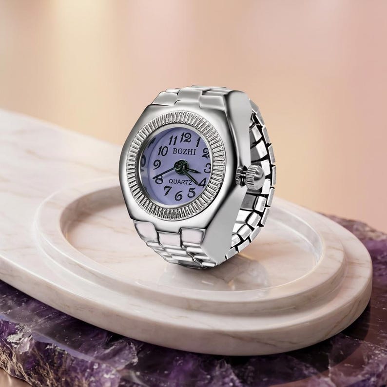 May include: A silver ring watch with a pale purple face and the word "BOZHI" printed on the watch face. The watch has a quartz movement and a flexible band. The watch is displayed on a white marble tray.