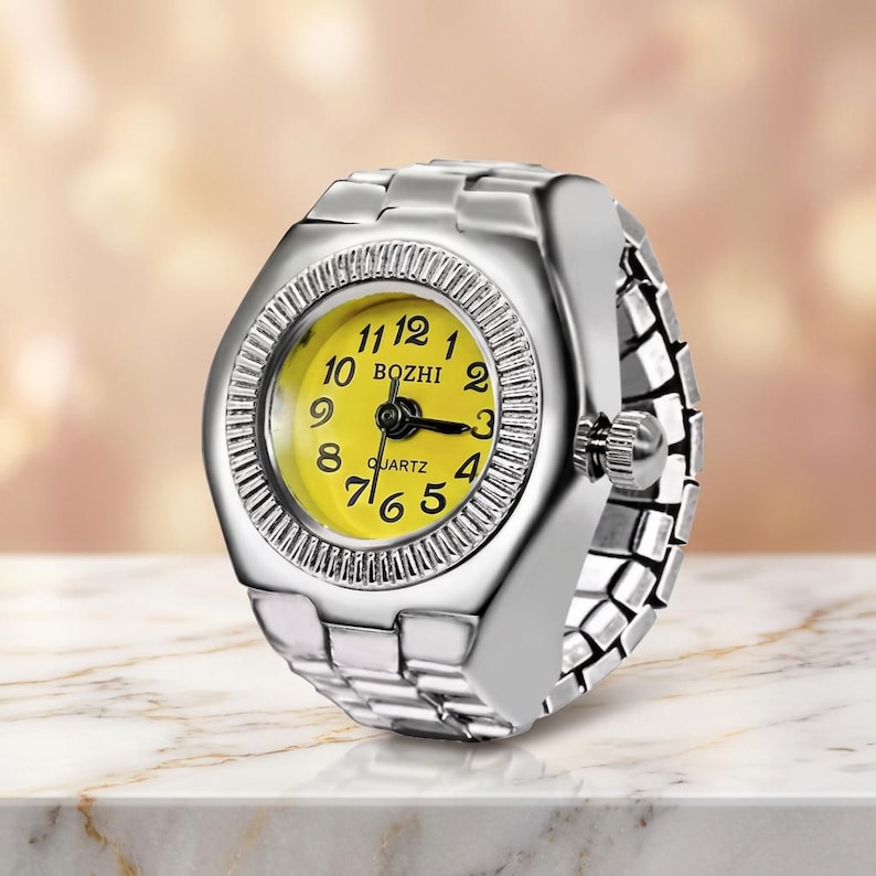 May include: A silver ring watch with a yellow face. The quartz watch has black hands and white numerals. The ring band is made of linked metal pieces. The watch face is surrounded by a textured bezel. The word "BOZHI" is printed on the watch face.