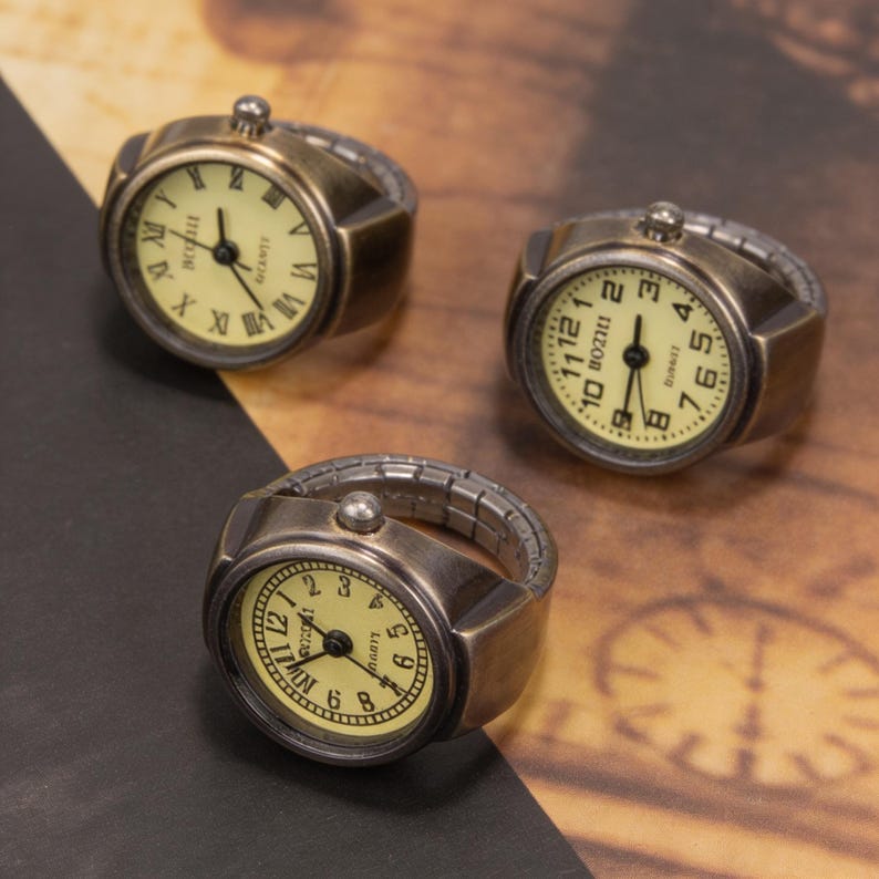 May include: Three antique-style ring watches with cream-colored faces and black hands. The rings have a bronze finish and feature Roman numeral hour markers. The watch bands are expandable.