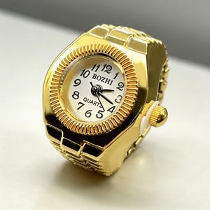 May include: A gold-colored ring watch with a white face and black numerals. The watch face has the word "BOZHI" and "QUARTZ" printed on it. The ring watch has a gold-colored band and a small crown on the side.