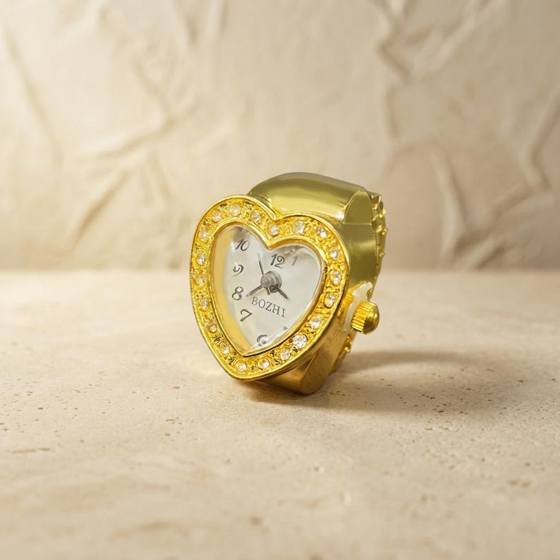 May include: A gold-toned ring watch with a heart-shaped face. The watch face is white with black numerals and hands, and the bezel is encrusted with small, clear crystals. The band is adjustable. The watch face has the word "BOZHI" printed on it.