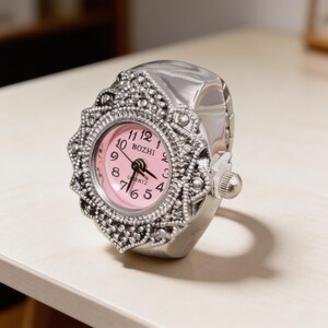 May include: A silver-toned ring watch with a pink face. The watch features an ornate, decorative bezel and a small crown for setting the time. The watch face displays the brand name "BOZHI".