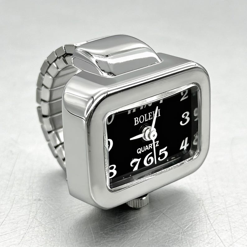 May include: A silver-toned ring watch with a rectangular face and a black dial. The watch face displays white numerals and hands, with the brand name "BOLEVI" visible. The band is a flexible, expandable design.