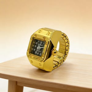 May include: A gold-colored ring watch with a square black watch face. The watch face displays white numerals and hands. The band is made of gold-colored, flexible links. The watch is resting on a light brown wooden surface.