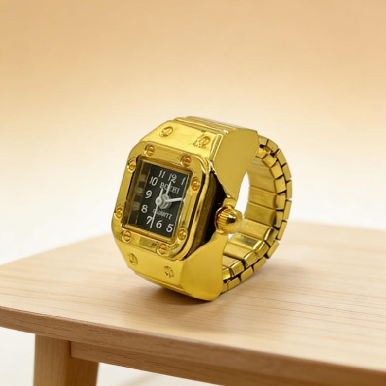 May include: A gold-colored ring watch with a square black watch face. The watch face displays white numerals and hands. The band is made of gold-colored, flexible links. The watch is resting on a light brown wooden surface.