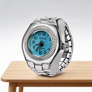 May include: A silver ring watch with a light blue face. The watch features a round face with black numerals and hands. The band is made of linked metal. The watch is on a light brown wooden surface.