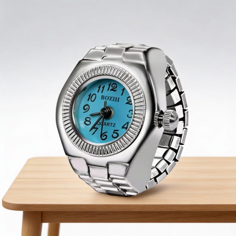 May include: A silver ring watch with a light blue face. The watch features a round face with black numerals and hands. The band is made of linked metal. The watch is on a light brown wooden surface.