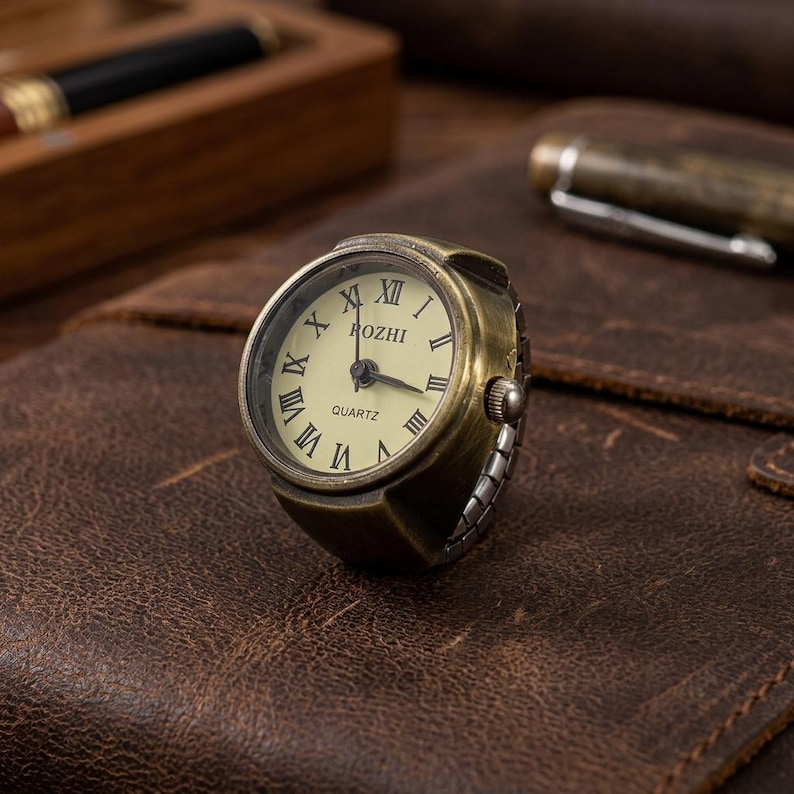 May include: Antique bronze-colored ring watch with a cream-colored face and black Roman numerals. The watch face reads "ROZHI QUARTZ". The ring watch is set on a brown leather surface.