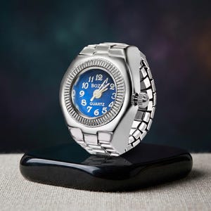 May include: A silver ring watch with a blue face and white numerals. The watch has a quartz movement and a linked metal band. The watch is displayed on a black, rectangular base.
