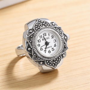 May include: A silver-toned ring watch with a white face and black numerals. The watch face is surrounded by an ornate, textured bezel. The ring band is a simple, polished metal.