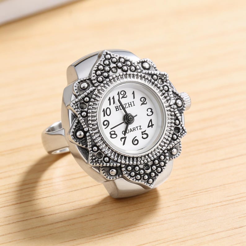 May include: A silver-toned ring watch with a white face and black numerals. The watch face is surrounded by an ornate, textured bezel. The ring band is a simple, polished metal.