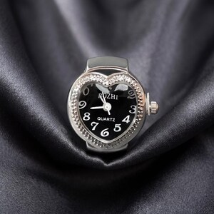 May include: A silver ring watch with a heart-shaped face. The black watch face has white numerals and hands, with the word "ROZHI" above the hands and "QUARTZ" below. The bezel is encrusted with small, clear crystals.