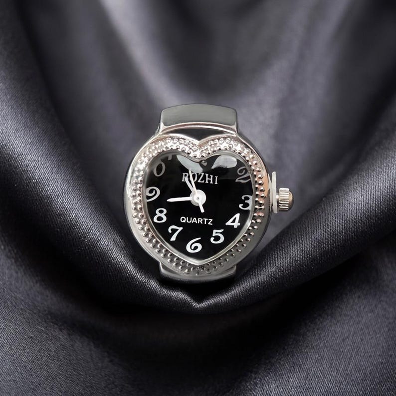 May include: A silver ring watch with a heart-shaped face. The black watch face has white numerals and hands, with the word "ROZHI" above the hands and "QUARTZ" below. The bezel is encrusted with small, clear crystals.