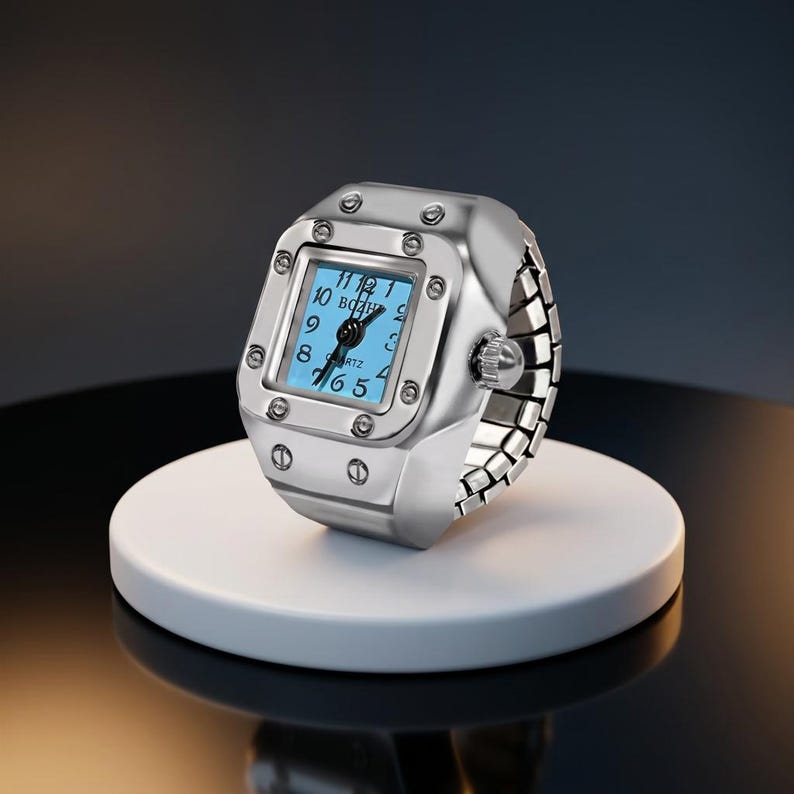 May include: A silver ring watch with a square face and a light blue dial. The watch has a quartz movement and a flexible band. The ring watch is displayed on a white circular stand.