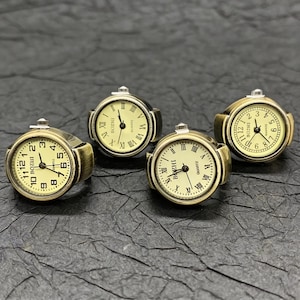 May include: Four antique-style ring watches with gold-toned metal bands and cream-colored faces. The watch faces display Roman numerals and the brand name "BOZHI". The rings are arranged on a dark, textured surface.
