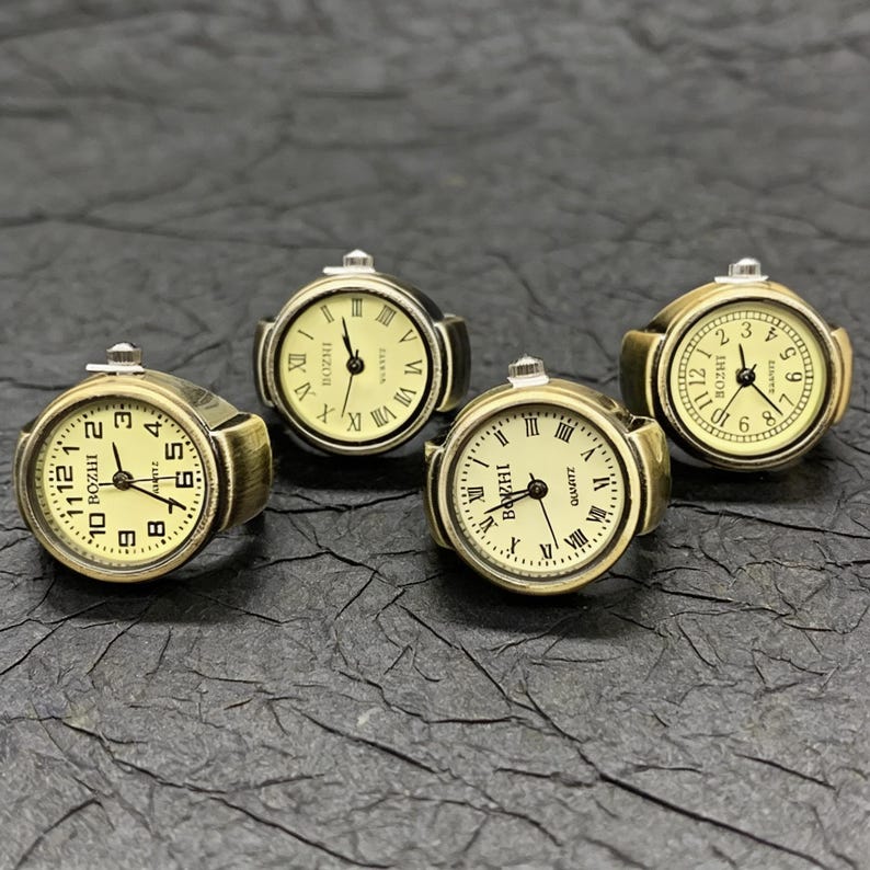 May include: Four antique-style ring watches with gold-toned metal bands and cream-colored faces. The watch faces display Roman numerals and the brand name "BOZHI". The rings are arranged on a dark, textured surface.