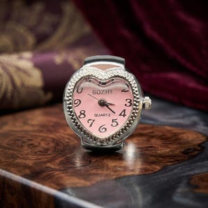 May include: A heart-shaped ring watch with a pink face and silver bezel. The watch face displays the numbers 3, 4, 5, 6, 7, 8, 9, and 0, with the brand name "BOZHI" at the top. The watch has a quartz movement.