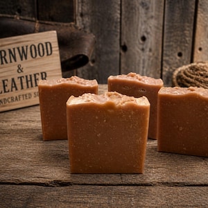 May include: Four square, light brown soaps with textured surfaces are arranged on a rustic wooden table. A wooden sign that reads "Barnwood & Leather Handcrafted Soap" is in the background. The soaps have a natural, handmade appearance.