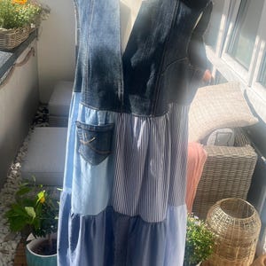 Könnte beinhalten: Ein Patchwork-Kleid auf einer Schaufensterpuppe, mit Jeans-, gestreiften und einfarbigen blauen Stoffen. Das ärmellose Kleid hat einen V-Ausschnitt und eine Tasche. Das Kleid ist eine Mischung aus hellen und dunklen Blautönen mit fließendem, gestuftem Design.