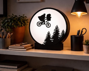 May include: A black and white circular lamp featuring a silhouette of a person and an alien riding a bicycle in front of a forest. The lamp has a black base and frame, and is illuminated from within. The lamp is on a shelf with other items.