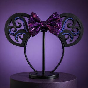 Peut inclure: Serre-tête noir avec des oreilles décoratives et un nœud papillon violet à paillettes. Les oreilles présentent un motif tourbillonnant avec des accents bleus. L'ensemble est posé sur un support noir, sur fond violet.